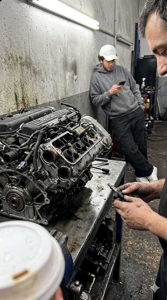Ferrari V8 Engine on Repair Shop Workbench – Candid iPhone Photo with Mechanic Hands and Man on Phone in Background — Portrait AI art prompt example for Gemini and Stable Diffusion